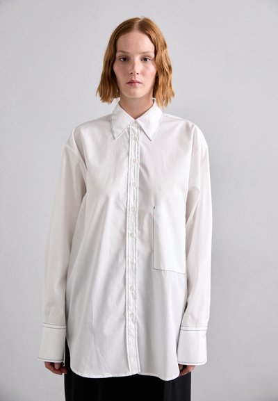 Chemise blanche à manches longues avec un devant à boutons, un col pointu et une poche unique. Présente des coutures contrastées et une coupe décontractée.