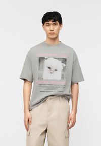 Young man wearing a gray oversized t-shirt with a missing white cat poster and beige cargo pants against a plain white background.