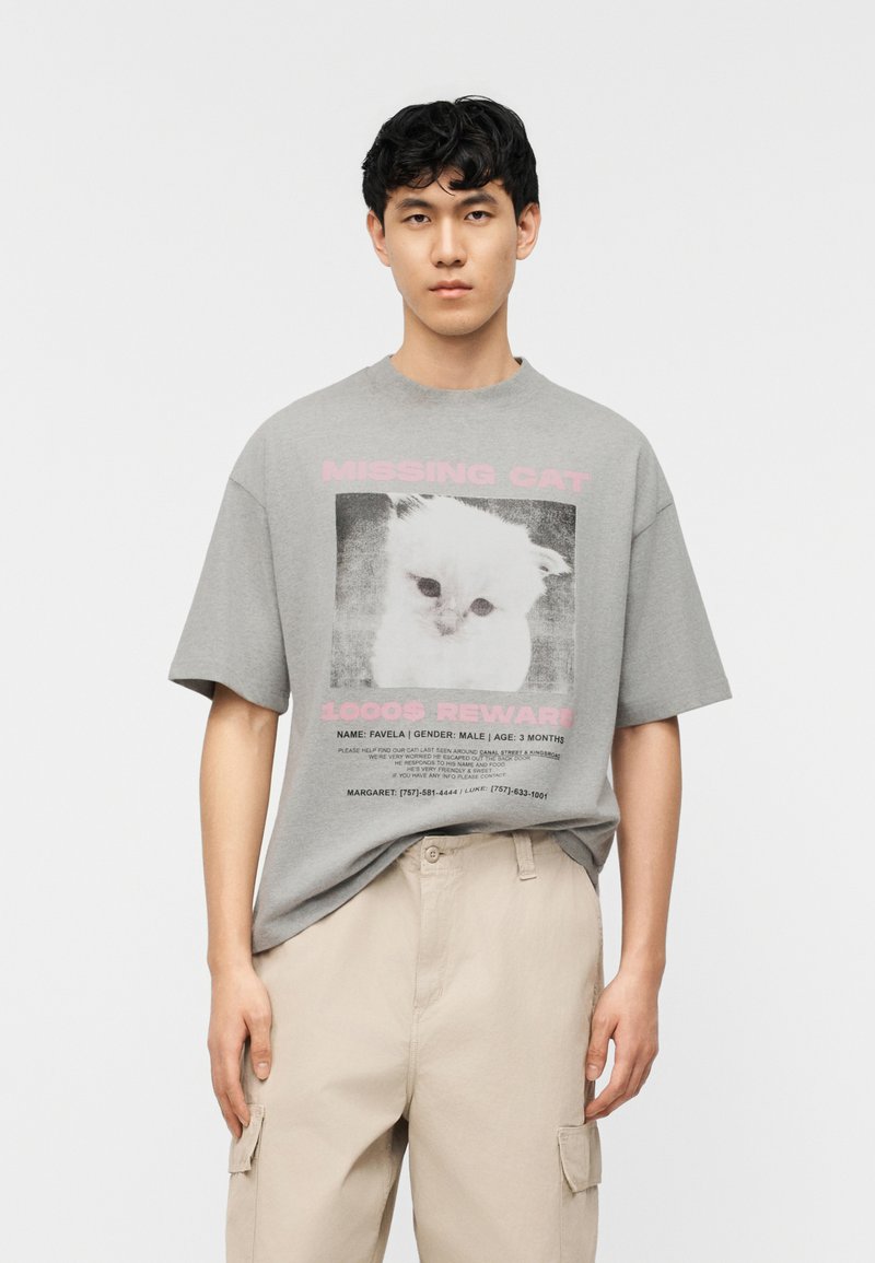 Young man wearing a gray oversized t-shirt with a missing white cat poster and beige cargo pants against a plain white background.