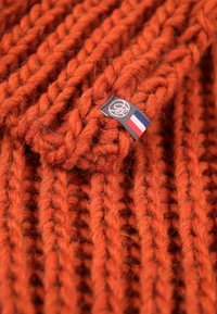Pull en maille épaisse d'un vibrant orange brûlé. Présente une texture en côtes prononcée et une petite étiquette avec un motif de drapeau attachée.