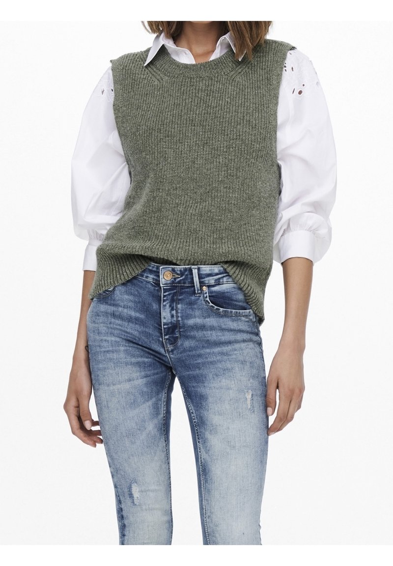 Pull sans manches vert tricoté avec un motif texturé, superposé sur une chemise blanche à manches bouffantes, associé à un jean bleu déchiré.
