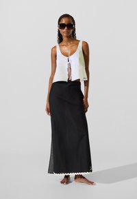 Ženska, ki nosi velike črne sončna očala, brezrokavnik z vozlom spredaj v beli barvi in dolgo črno krilo z belo valovito obrobo, stoji bosa.