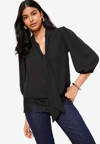 Donna che indossa una blusa nera con scollatura a chiave, maniche a sbuffo, orlo in pizzo, jeans blu scuro, orecchini a cerchio dorati e braccialetto argento, in posa con la mano sui fianchi.