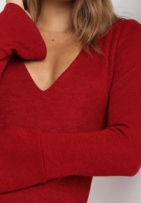 Röd stickad tröja med djup V-ringning och långa ärmar med en slät textur. Tyget har ett subtilt, fint mönster.