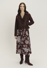 Giacca corta in finta suede bordeaux con chiusura a zip, abbinata a una gonna lunga a fiori in tonalità simili e stivali bordeaux al ginocchio.
