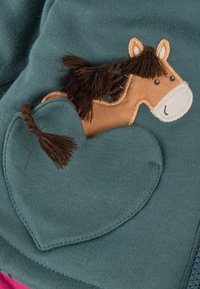Tessuto color teal con un design ricamato di un cavallo marrone e un cuore. Il cavallo ha una criniera in filato scuro. Texture morbida, stile casual.