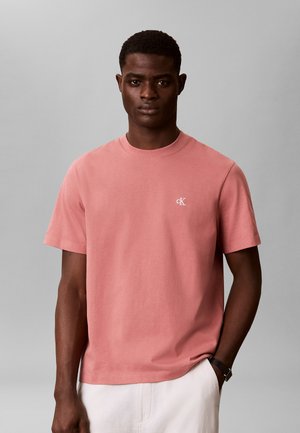 Ung mand iført en lakserød Calvin Klein t-shirt og hvide bukser, stående foran en ensfarvet grå baggrund.