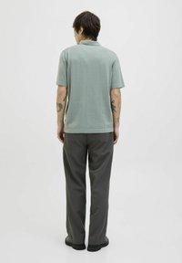 Polo shirt à manches courtes de couleur vert clair associé à un pantalon ample gris foncé. La chemise présente une texture lisse et la tenue est complétée par des chaussures noires.