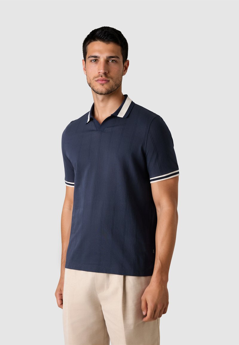 Mann steht, trägt ein marineblaues Polohemd mit kurzen Ärmeln und weißer Einfassung an Kragen und Ärmeln sowie eine hellbeige Hose, neutraler Hintergrund.