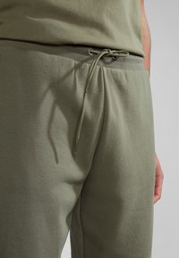 Bavlněné tepláky olivově zelené barvy s nastavitelným pasem na stahovací šňůrku, mají měkkou texturu a elastický pas, na látce jsou vidět záhyby a mírné vrásky.