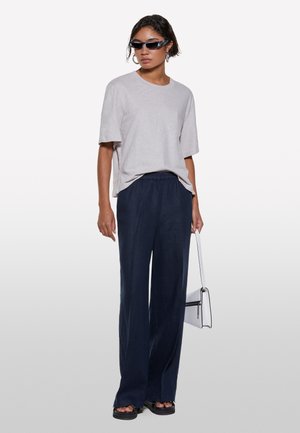 T-shirt oversize grigio chiaro, pantaloni a gamba larga blu navy, sandali platform neri, borsa bianca. Occhiali da sole con una montatura sottile completano l'outfit.