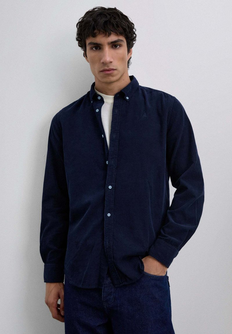 Camisa de botones azul marino, mezcla de algodón, con cuello extendido, mangas largas y botones de color azul claro. Llevada sobre una camiseta blanca.