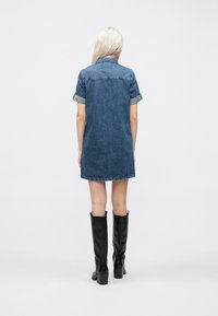 Mørkeblå denimkjole. Har rullede korte ermer, krage og en frynset kant. Brukt med svarte knelengde støvler.