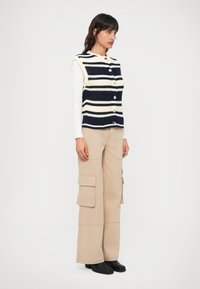 Colete de malha riscas azul-marinho e creme com decote redondo e botões à frente, combinado com calças de carga bege e botas de tornozelo pretas.