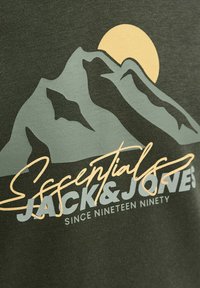 Donkergroene shirt met een bergafbeelding in lichtgroen en een gele zon. De tekst luidt "Essentials Jack & Jones" in wit en geel.