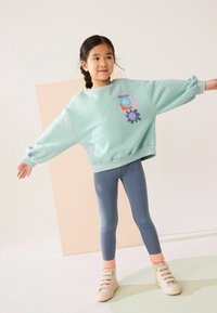 Menta zöld, oversized pulóver színes virágmintákkal, kék leggings-szel és bézs, műszőrmével díszített magasszárú cipőkkel párosítva.