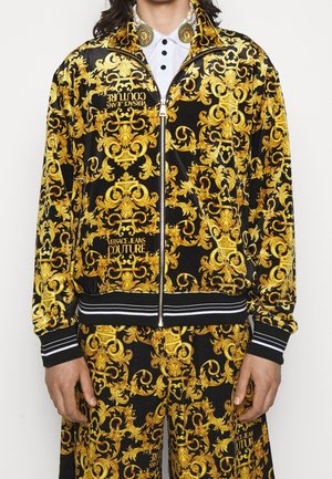 Schwarz-goldene, verzierte Barockjacke und passende Hose mit Versace Jeans Couture Logos, getragen über einem weißen Hemd mit Kragen.