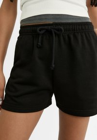 Kvinde iført sorte elastiske shorts med snor i taljen og en lagdelt hvid og grå ribbet top, stående mod en hvid baggrund.