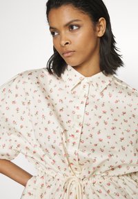 Camisa floral em tecido creme com pequenos padrões de rosas cor-de-rosa. Apresenta um colarinho abotoado e cintura com cordão para ajuste. Textura suave.