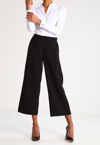 Weißes Hemd mit langen Ärmeln und Kragen, kombiniert mit hochtaillierten, weit geschnittenen schwarzen Hosen und glänzenden schwarzen Absätzen.