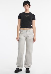 Maglietta nera corta con logo "GUESS" dorato; jeans grigi chiari a gamba larga; stivali neri con lacci; sfondo bianco semplice.