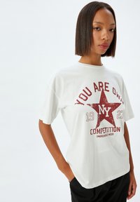 Camiseta blanca de algodón con un gráfico de una estrella roja y el texto "ERES PROPIEDAD DE NY 1922 COMPETITION UNBREAKABLE MOOD." Corte ancho con mangas cortas.