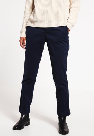 Personne portant un pantalon droit bleu marine foncé, un pull crème en tricot et des bottines noires, debout, une main dans la poche.