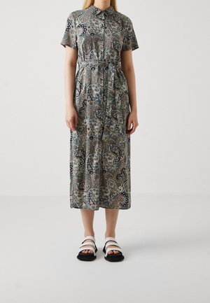 Femme portant une robe midi boutonnée et ceinturée avec des motifs cachemire et des manches courtes, associée à des sandales blanches et noires, debout sur un sol blanc.