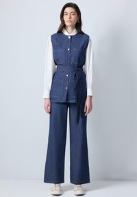 Gilet en denim avec quatre poches, fermeture à boutons et ceinture à la taille, associé à un pantalon en denim à jambes larges. Les deux sont de couleur bleu foncé. Chemise blanche en dessous.