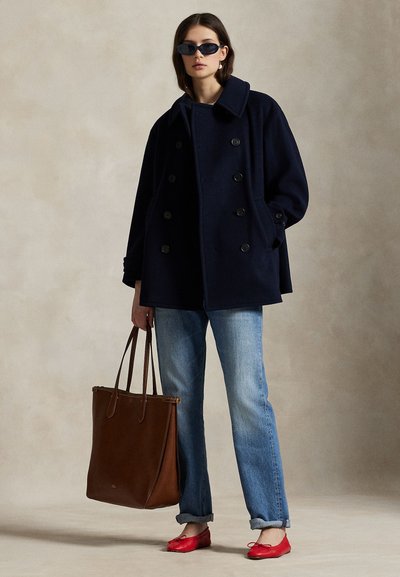 Naine kannab tumesinist kahekordsete nööpidega mantlit, päikeseprille, siniseid teksaseid, punaseid lameda tallaga kingi ja hoiab suurt pruuni nahast käekotti ühevärvilise tausta ees.