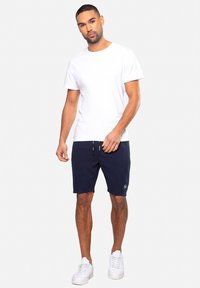 Weißes T-Shirt mit kurzen Ärmeln und navyblauen Shorts mit Kordelzug. Das Modell trägt weiße Sneaker. Die Shorts verfügen über ein dezentes Streifenmuster und ein Logodetail.