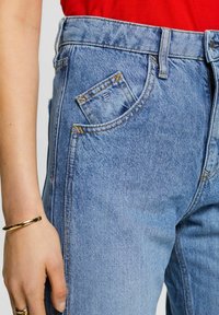 Helle blaue Jeans mit hoher Taille, ausgestattet mit einer kleinen Fronttasche, sichtbaren Nähten und einer weichen, strukturierten Oberfläche.