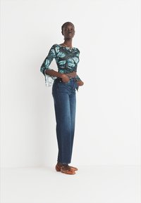 Modra majica z dolgimi rokavi z vzorcem metuljev, temno modri jeans z visokim pasom in rjave pletene sandale, na belem ozadju.