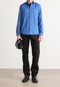 Camicia blu a maniche lunghe con bottoni, pantaloni neri, che tiene una piccola borsa nera. Tessuto morbido, vestibilità su misura e calzature nere. Design minimalista.