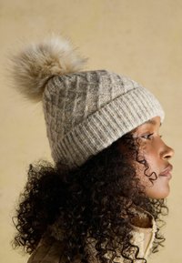 Knitted beige beanie with a textured diamond pattern and a light faux fur pom-pom on top. Ribbed band at the base for a snug fit.