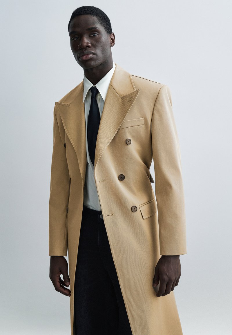 Abrigo cruzado color beige con solapas afiladas, tela texturizada, bolsillos laterales y botones; combinado con una camisa clara y corbata negra.