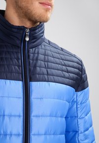 Fodrad jacka med dragkedja, navyblå övre del och ljusblå nedre del, strukturerad kviltad design och hög krage.