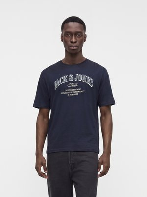 Ung mand stående med fronten fremad iført en marineblå Jack & Jones T-shirt med print og mørke jeans mod en ensfarvet lys baggrund.