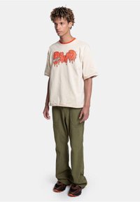 Beige oversized t-skjorte med rød krage og oransje dryppende tekst, kombinert med grønne baggy bukser og svarte joggesko med oransje detaljer.