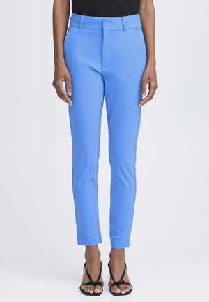 Personne portant un pantalon bleu ciel ajusté longueur cheville, un haut noir sans manches et des sandales à talons noires, debout sur un fond uni.