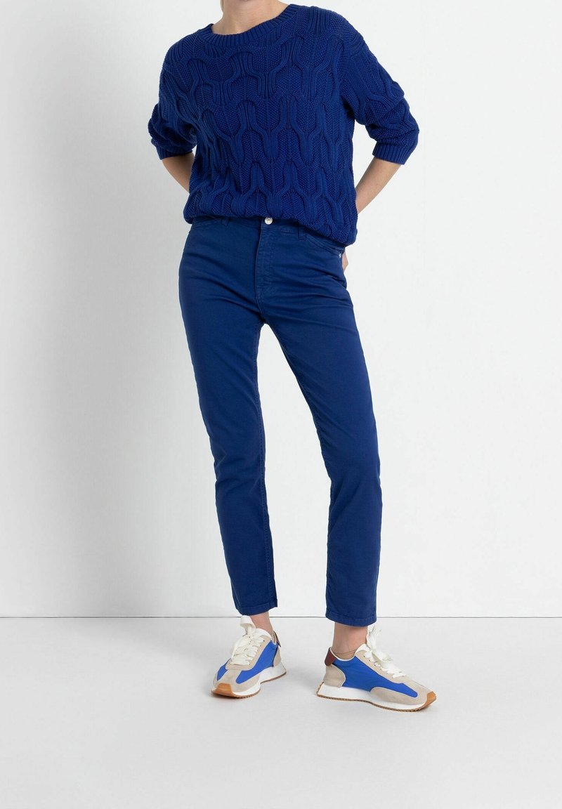 Pull bleu royal en maille torsadée à la coupe décontractée, associé à un pantalon slim bleu assorti et des baskets blanches avec des accents bleus et beige.