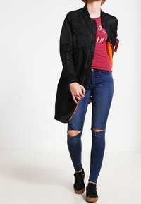 Czarna kurtka bomberka z pomarańczową podszewką, noszona na czerwonej koszuli i niebieskich dżinsach z przetarciami. Czarne buty o teksturowanej podeszwie dopełniają całość.