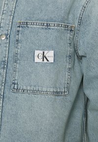 Světle modrá džínová košile s náprsní kapsou. Na levé náprsní kapse je bílá visačka s nápisem "Calvin Klein Jeans" a černým logem.