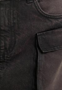 Černá džínsovina s texturovaným povrchem, přední kapsou a pečlivě šitými okraji. Design je utilitární s úzkým střihem.