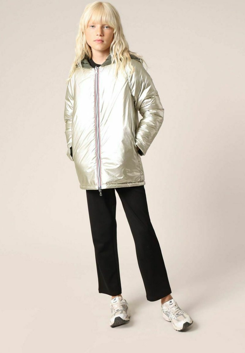 Veste en duvet argentée, surdimensionnée, avec un col haut et une fermeture éclair contrastante. Associée à un pantalon noir et des baskets blanches. Texture lisse.