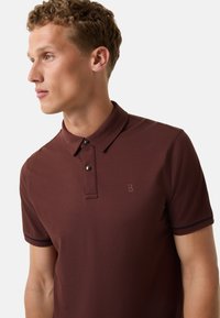 Burgundské polo tričko s klasickým límečkem, zapínáním na dva knoflíky, jemným logem a kontrastními vroubkovanými manžetami. Hladká, strukturovaná tkanina.