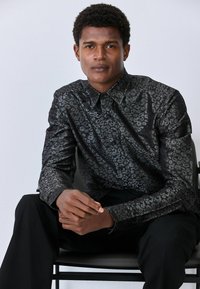 Czarna koszula z guzikami, z teksturowanym wzorem w lamparcie cętki, długie rękawy i kołnierz, noszona przez siedzącą osobę na neutralnym tle.