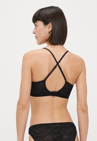 Črni modrček z nastavljivimi križnimi naramnicami, teksturirana tkanina z diskretnim vzorcem in zaponko na hrbtni strani.