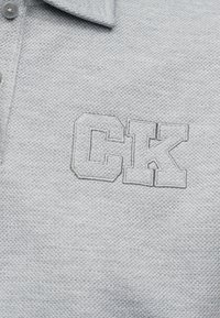 Tricou polo gri deschis texturat, cu guler și nasturi, având litere brodate în relief "CK" pe piept.