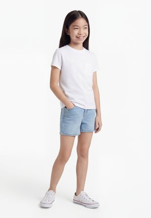 Glimlachend meisje met lang donker haar draagt een wit T-shirt, lichtblauwe spijkershorts en witte sneakers, staande met één hand in de zak.